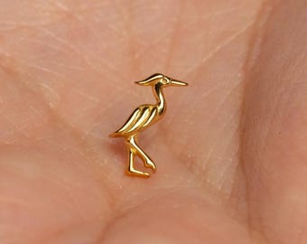 May include: A small, gold-colored metal charm shaped like a standing heron or crane bird. The charm features a smooth, reflective surface with a pointed beak, a slender neck, and detailed wing markings, captured in a side profile view.