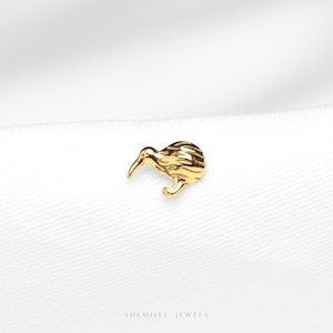 Peut inclure: Une petite broche dorée en forme de kiwi. La broche représente l'oiseau sans ailes avec un long bec et des plumes texturées. La broche est posée sur un fond blanc. Le nom de la marque 'SHEMISLI JEWELS' est en bas.