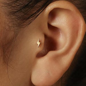 May include: A small, gold-coloured lightning bolt stud earring is shown in the ear. The earring is a simple, minimalist design, and the gold colour provides a bright contrast against the skin. The earring is a stylish accessory.