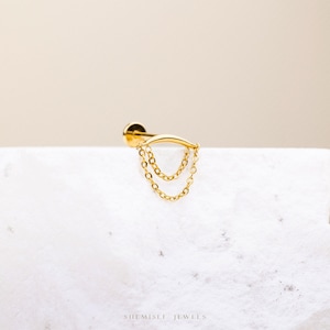 May include: A gold earring featuring a curved bar design with two hanging chain strands. The earring has a small, round stud backing. The jewellery is displayed on a white surface with the text "SHEMISLI JEWELS" at the bottom.