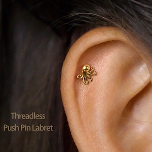 May include: A gold-coloured, threadless push pin labret earring shaped like an octopus. The small earring has a rounded head and eight curled tentacles. The text "Threadless Push Pin Labret" is visible.