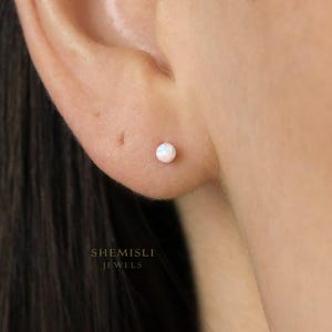 Peut inclure: Gros plan sur une petite boucle d'oreille ronde en opale. La boucle d'oreille est fixée dans le lobe de l'oreille d'une personne. L'opale a un aspect blanc et irisé. Le nom de la marque "SHEMISLI JEWELS" est imprimé en blanc en bas de l'image.