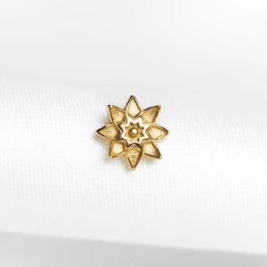 Peut inclure: Un bijou doré en forme d'étoile à huit branches. Le centre et les bords sont remplis d'émail blanc, créant un design contrasté. Le bijou est posé sur un fond blanc.