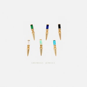 May include: Gold-coloured earrings with a spike design, each topped with a different coloured rectangular gemstone: green, blue, black, clear, opal, and turquoise. The earrings are displayed on a white background. The text "SHEMISLI JEWELS" is at the bottom.