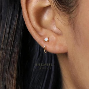 Peut inclure: Gros plan sur une oreille avec deux boucles d'oreilles. L'une est une petite puce d'oreille en or avec une opale blanche ronde. L'autre est un petit anneau d'oreille en or. Le texte "SHEMISLI JEWELS" est visible.
