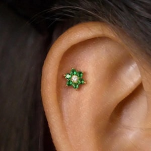 May include: A gold-coloured flower-shaped earring featuring green gemstones and a central white gemstone. The earring is set against the backdrop of an ear, highlighting its design and placement.