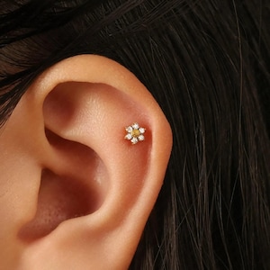 Peut inclure: Boucle d'oreille en forme de fleur dorée avec un centre jaune et des pétales en pierres précieuses blanches. La boucle d'oreille est placée dans l'oreille d'une personne, avec des cheveux foncés visibles en arrière-plan. Bijou délicat.