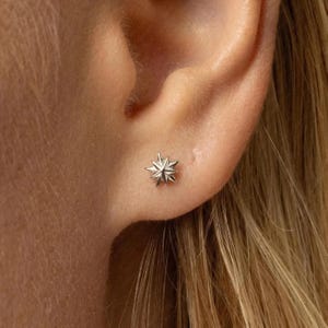 May include: A small, silver starburst stud earring is shown on an ear. The earring has a detailed, multi-pointed design. The ear is against a background of blonde hair.