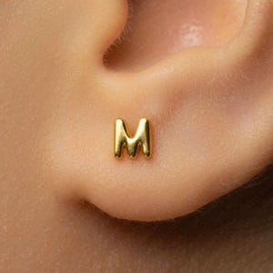 May include: A small, polished gold-tone stud earring shaped like the capital letter M. The earring features a smooth, rounded surface and a metallic finish, designed to be worn as a single piece in the earlobe with a simple, clean aesthetic.