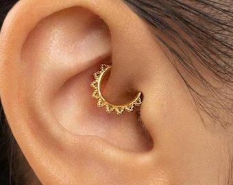 Può includere: Primo piano di un anello per setto nasale dorato con cerniera e design a forma di sole. Il gioiello è indossato in un orecchio forato. Un piccolo orecchino a bottone dorato è visibile anche nel lobo.