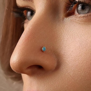 Peut inclure: Gros plan sur un nez orné d'un piercing en or. Le bijou présente une pierre turquoise en forme de goutte d'eau sertie dans une lunette en or. La peau autour du nez est lisse et a un ton naturel.