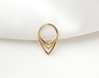 Pode incluir: Um piercing de septo dourado com um design único em forma de gota. O piercing apresenta um topo circular e uma base pontiaguda, com duas camadas em forma de seta. As bordas são adornadas com pequenas contas espaçadas, adicionando textura e detalhes à joia.
