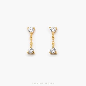 May include: A pair of gold drop earrings, each with a clear, triangular gemstone. The earrings have a small gemstone at the top, connected by a chain to a larger gemstone below. The earrings are set against a white background. The brand name is visible.