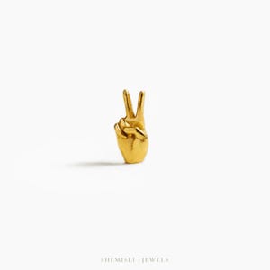 May include: A gold-coloured metal sculpture of a hand forming the peace sign. The sculpture has detailed fingers and a textured surface. The background is plain white, emphasising the sculpture's form. The text "SHEMISLI JEWELS" is at the bottom.