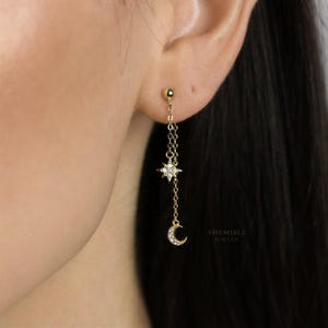 May include: Gold-coloured celestial earrings with a star and crescent moon design. The earrings hang from a small ball stud, with a star-shaped charm and a crescent moon charm. The earrings are attached by delicate chains.