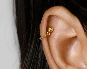 Puede incluir: Un piercing de oreja dorado. Un pequeño perno de bola está conectado por una delicada cadena a una segunda bola. La joya se coloca sobre el fondo de una oreja y cabello oscuro.