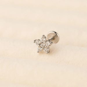 May include: A silver stud earring with a star-shaped design. The star is made up of six clear, round gemstones. The earring post and backing are also silver. The background is a soft, neutral colour.