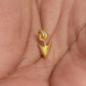 Peut inclure: Un petit pendentif en forme de tulipe de couleur or. Le pendentif présente une tête de fleur détaillée et une tige avec deux feuilles pointues. Le bijou est tenu dans une main, mettant en valeur sa taille et son design.