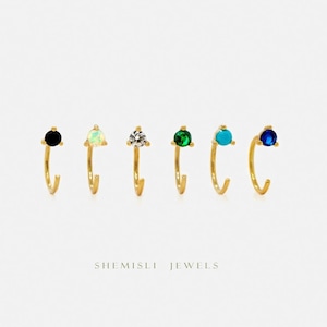 May include: Six gold-tone hoop earrings, each with a different gemstone: black, opal, clear, green, turquoise, and blue. The earrings are displayed in a row against a white backdrop. The brand name "SHEMISLI JEWELS" is printed below the earrings.