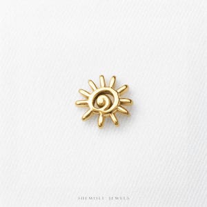 May include: A small gold-toned sun-shaped charm with a central spiral design and radiating rays. The piece features a polished metallic finish and is positioned above the text Shemisli Jewels printed in a simple black sans-serif font.