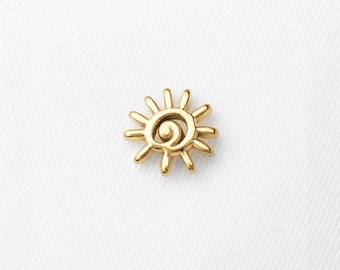 May include: A small gold-toned sun-shaped charm with a central spiral design and radiating rays. The piece features a polished metallic finish and is positioned above the text Shemisli Jewels printed in a simple black sans-serif font.