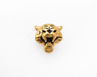 Könnte beinhalten: Ein goldfarbener Metallanhänger in Form eines brüllenden Tigerkopfes mit schwarzer Emaillierung an Augen, Nase und Maul. Der Tiger zeigt modellierte Ohren, Schnurrhaare und gefletschte Zähne. Der Text SHEMISLI JEWELS ist unten aufgedruckt.