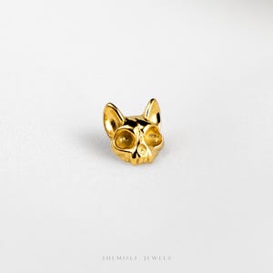 Peut inclure: Une petite boucle d'oreille clou en métal doré en forme de crâne de chat avec des oreilles pointues et des orbites creuses. La pièce présente une surface lisse et réfléchissante. Le texte SHEMISLI JEWELS est inscrit en bas de l'image.