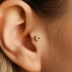 May include: A close-up of a gold star-shaped earring with a small, clear gemstone at its centre. The earring is set against the skin of an ear, showcasing its intricate design and sparkle. The earring is a stud style, perfect for everyday wear.