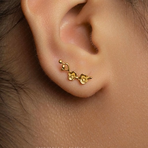 May include: A gold ear cuff with a floral design. The cuff curves along the ear, with three flower-shaped elements and a delicate stem. The jewelry is worn on the ear, showcasing its intricate details and elegant style.