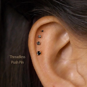 May include: An ear with four black stud earrings of varying sizes. The earrings are set in gold. The text "Threadless Push Pin" is visible on the left side of the image.