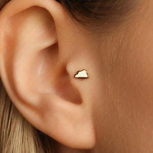 May include: A small, gold-coloured cloud-shaped earring. The earring is a stud style, with a white enamel filling the cloud shape. The earring is shown in a close-up view, worn on an ear.