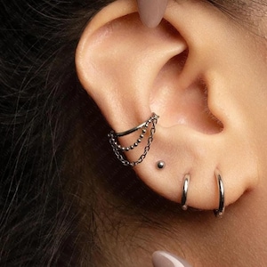 May include: An ear featuring multiple pieces of jewellery. A silver ear cuff with three hanging chains is visible, alongside a small stud and two silver hoop earrings. The jewellery is set against the backdrop of the ear and skin.