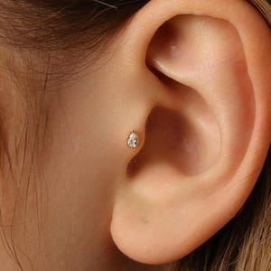 Peut inclure: Gros plan d'une boucle d'oreille dorée en forme de goutte d'eau avec une pierre claire et étincelante. La boucle d'oreille est sertie dans un cadre doré délicat et est positionnée sur l'oreille. L'oreille a un teint clair et lisse.