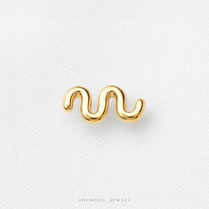 May include: A gold-toned metal earring shaped like a smooth, continuous wavy line with rounded ends. The piece features a polished, reflective surface and a curved, serpentine design. The text SHEMISLI JEWELS is printed at the bottom.