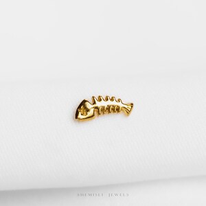 May include: A small, gold-coloured fish skeleton charm. The charm has detailed ribs and a head. The jewellery piece is set against a white background. The text "SHEMISLI JEWELS" is at the bottom of the image.