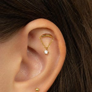 May include: A gold-coloured ear piercing featuring a small chain and a dangling, round, clear gemstone. The piercing is attached to the ear with a curved bar. The ear has a small gold stud piercing at the bottom.