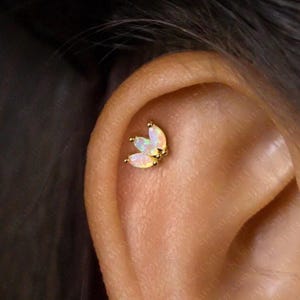 Peut inclure: Un piercing d'oreille doré avec trois pierres opalescentes en forme de marquise, disposées en motif floral. De petites perles dorées forment le centre, créant un groupe d'accents scintillants. Le bijou est placé sur l'oreille.