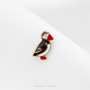 May include: A gold-toned enamel pin shaped like a puffin, featuring black, white, and red enamel fill. The bird is depicted in profile with a red beak and red feet. The text "SHEMISLI JEWELS" is printed in small black letters at the bottom.