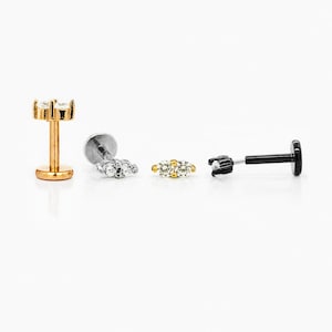 May include: Four pieces of jewellery are displayed against a white background. There are three labret studs with different designs and one gold-coloured stud with three clear gems. The other studs have clear and gold gems and a black post.