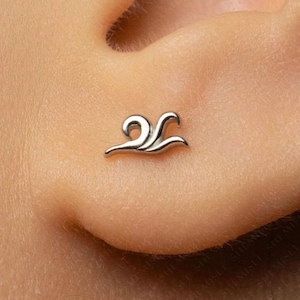 May include: A silver-toned metal stud earring shaped like a stylised wave or flame with a smooth, polished surface. The jewellery piece features a curved, flowing design that tapers to a point, securely positioned against the skin of an earlobe.