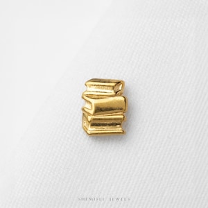 May include: A small gold-coloured charm shaped like a stack of three books. The piece features a polished metallic finish with defined horizontal lines representing the pages and covers of the books. The text SHEMISLI JEWELS is printed below.