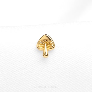 May include: A small gold-coloured mushroom stud earring with a polished finish. The cap features detailed, etched radial lines radiating from the centre stem. The text "SHEMISLI JEWELS" is printed in small, dark letters at the bottom of the frame.