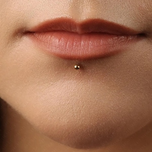 May include: Close-up of a person's lower lip with a gold-coloured stud piercing. The lips are a natural shade of pink and brown. The skin around the mouth is smooth and light-toned.