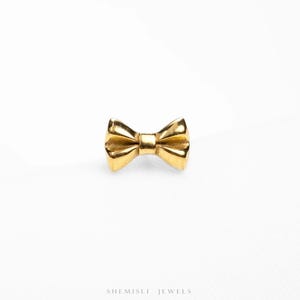 May include: A small, gold-coloured bow tie shaped jewellery piece. The bow tie is centered and has a polished, reflective surface. The jewellery is set against a white background. The text "SHEMISLI JEWELS" is at the bottom.