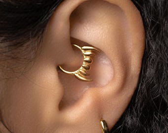Könnte beinhalten: Ein goldfarbener Ohrpiercingschmuck in Form eines Halbmonds mit klauenartigen Details. Der Schmuck wird im Knorpel des Ohrs getragen. Ein kleiner goldener Creolen-Ohrring und ein goldener Perlenohrring sind ebenfalls sichtbar.
