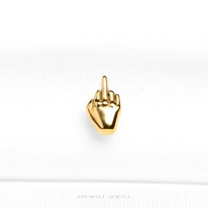 May include: A gold-coloured pendant in the shape of a hand giving the middle finger. The pendant is highly detailed and appears to be made of metal. The background is a plain white surface. The text "SHE MISLI JEWELS" is at the bottom.