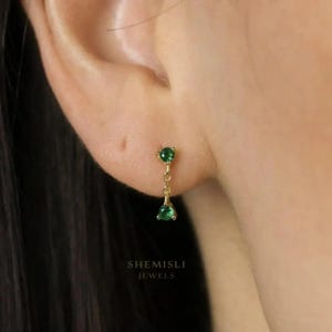 Peut inclure: Gros plan d'une boucle d'oreille dorée ornée de deux pierres vertes. La pierre supérieure est ronde, la pierre inférieure en forme de goutte, reliées par une fine chaîne. Le texte "SHEMISLI JEWELS" est visible.