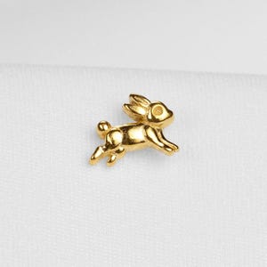 May include: A small, gold-coloured bunny figurine in a running pose. The bunny has detailed features, including ears, eyes, and paws. The figurine is set against a white background.