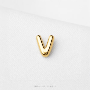 May include: A polished gold-tone metal charm in the shape of the letter V. The piece features a smooth, rounded surface with a reflective finish. The text SHEMISLI JEWELS is printed in small, dark letters at the bottom centre of the image.