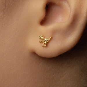 May include: A gold-coloured Tyrannosaurus Rex stud earring. The small dinosaur is detailed with visible ribs and a textured surface, and is affixed to an ear. The earring is a small, detailed piece of jewellery.
